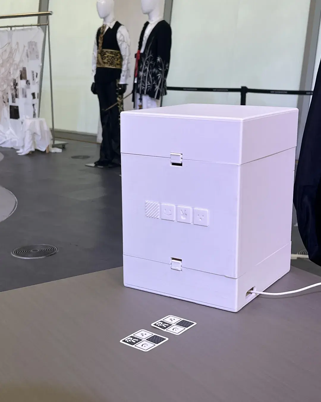 A white photo booth on the exhibition floor with its power cable on the ground, mannequins and costumes behind it
