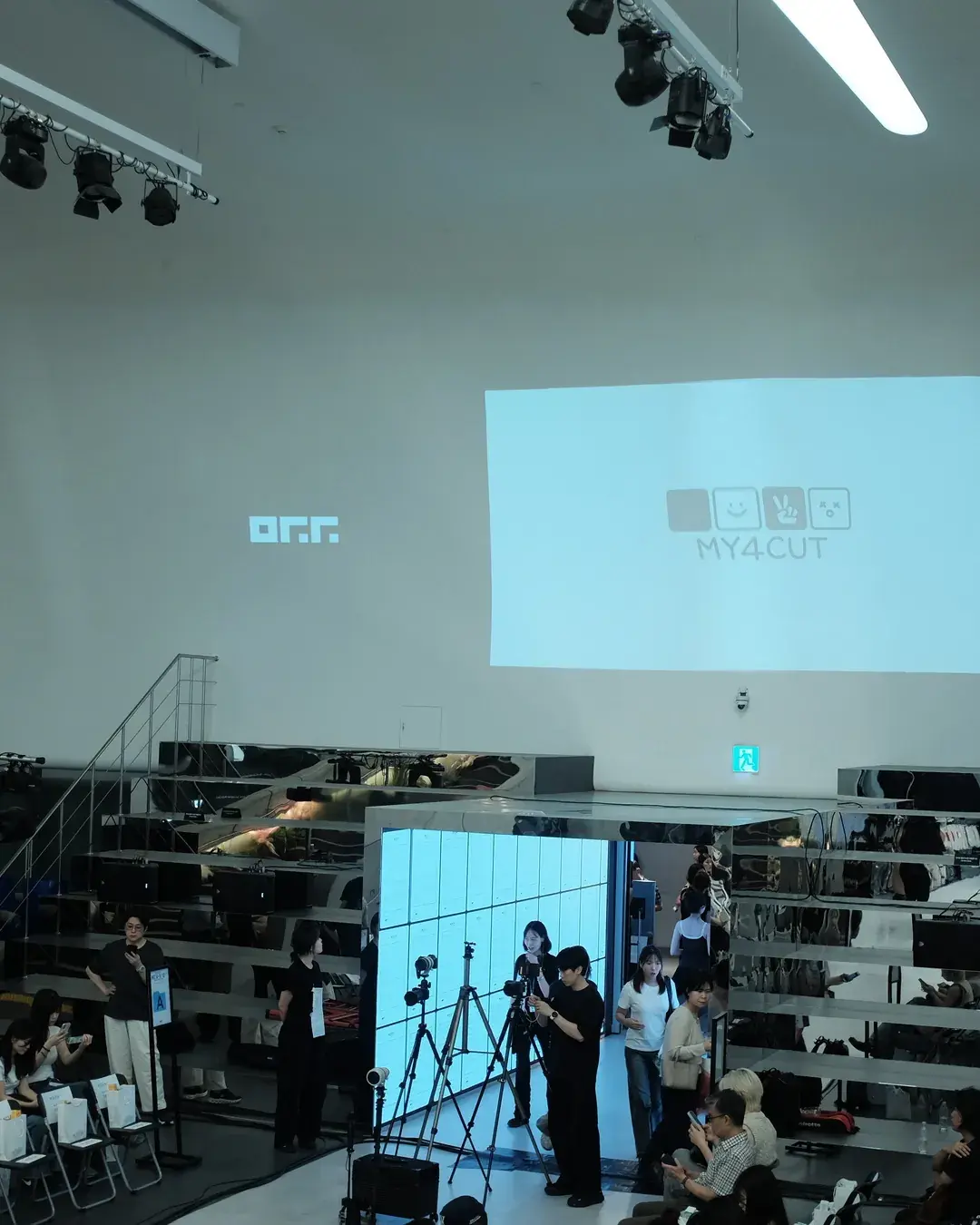 A stage screen at the DDP fashion show showing the O.F.F. and MY4CUT logos, with stage lights, seats and people filming