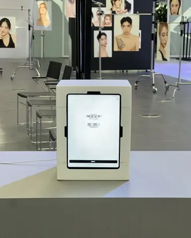 A small white photo booth on a stand in the DDP fashion show hall, with model photo panels and chairs behind it