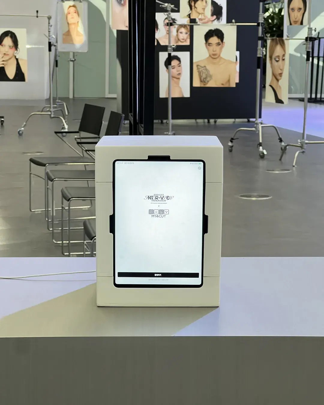 A small white photo booth on a stand in the DDP fashion show hall, with model photo panels and chairs behind it
