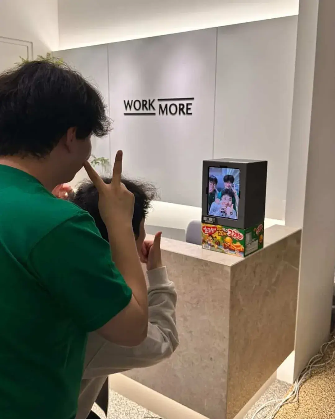 A small photo booth on a marble counter in front of a white WORKMORE wall, with two attendees from behind making V signs
