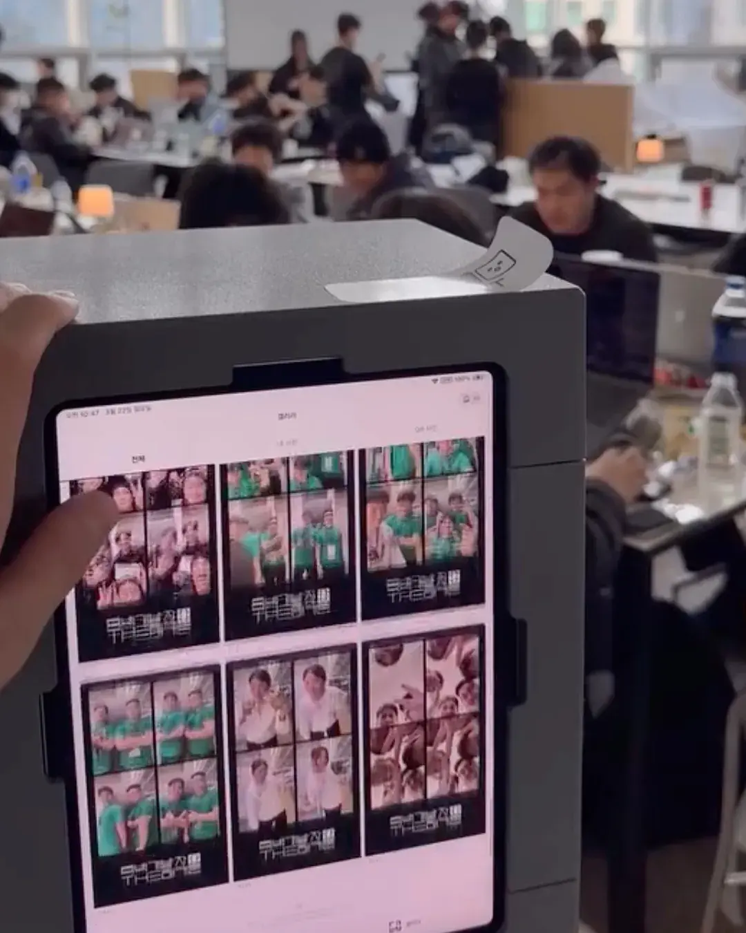 The booth iPad's gallery showing many 4-cut strips in a grid, one per team, with participants at laptops behind it