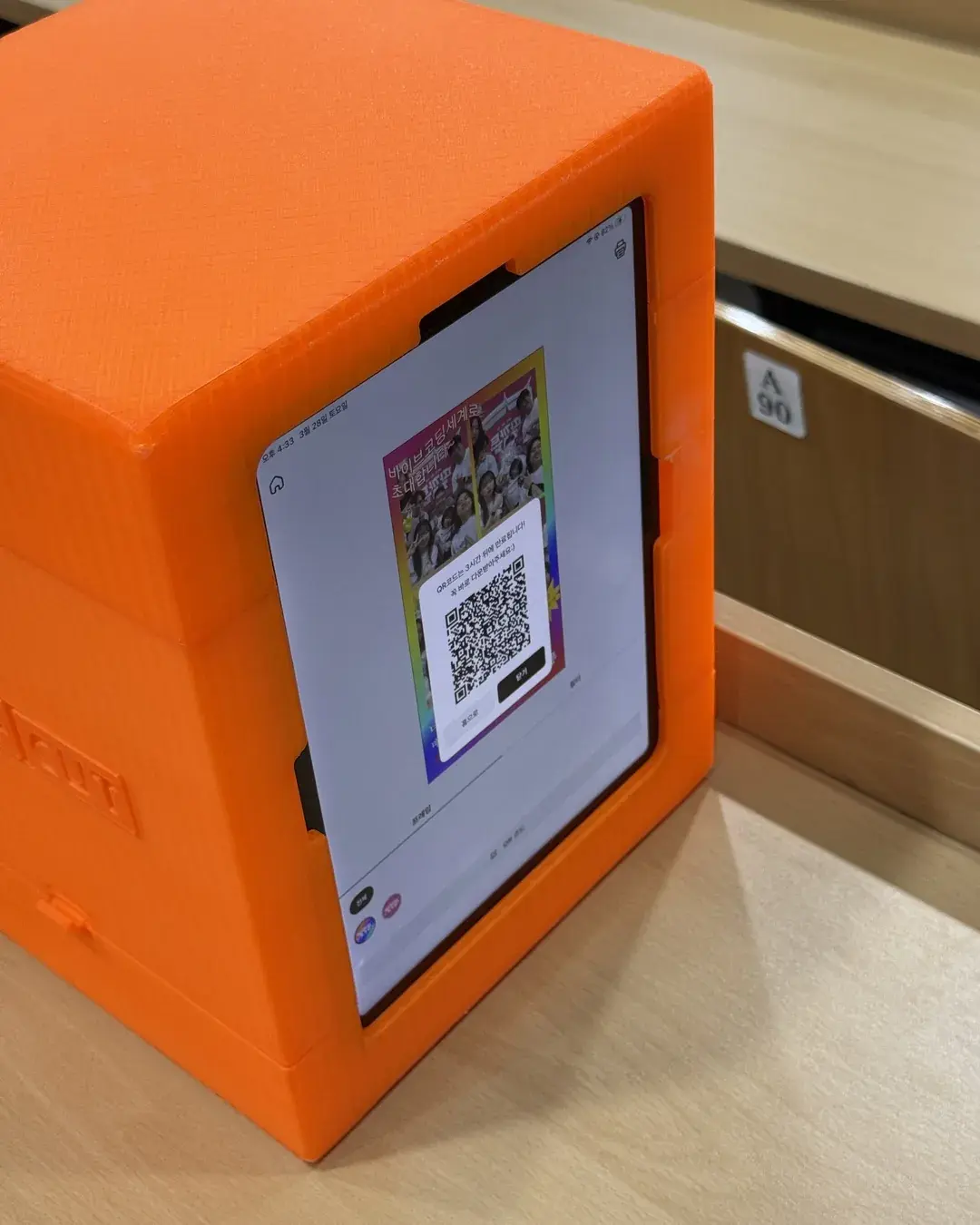 An orange mini booth on a table at Ddalkkakthon with the iPad showing the finished strip and a QR code