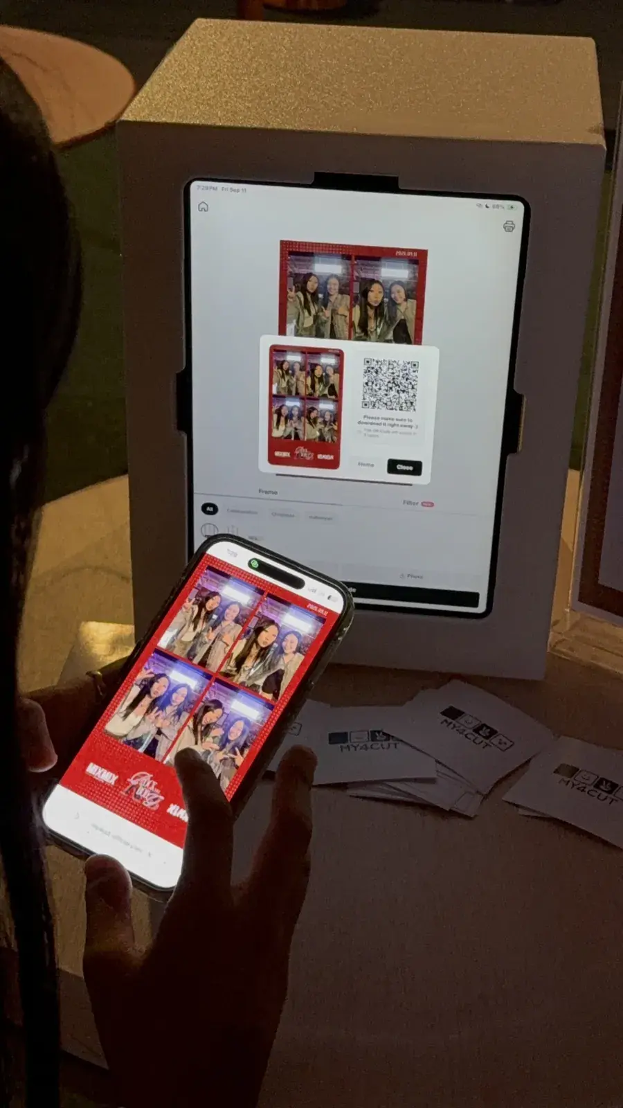 An iPad screen showing a finished red-frame photo and a QR code, with a phone in a hand showing the same photo saved