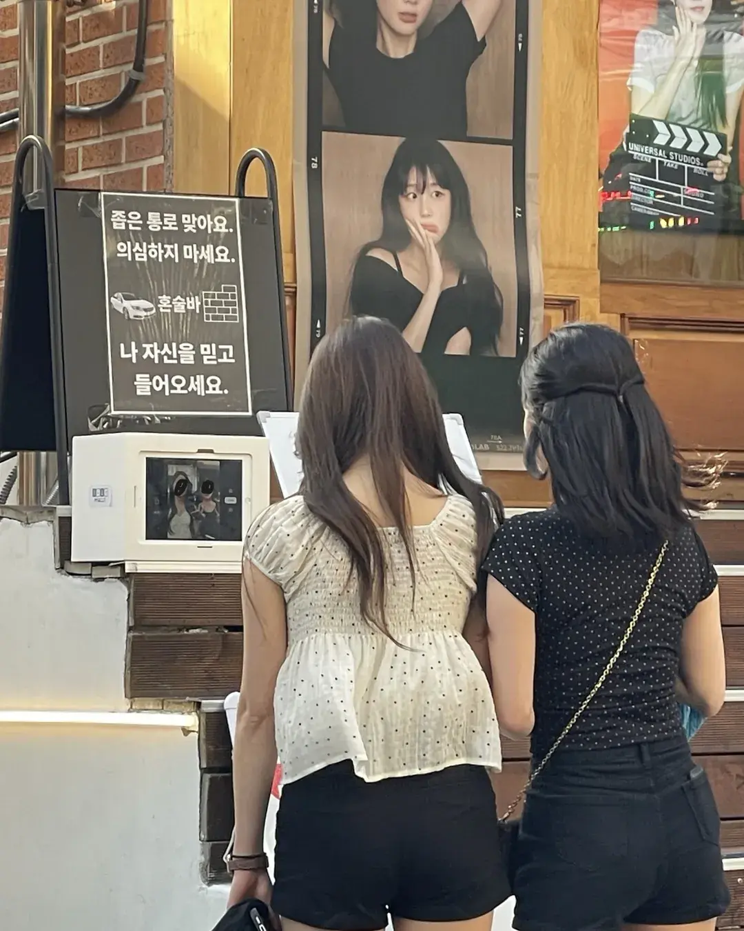 Two people seen from behind looking at a photo booth screen in a Haebangchon alley, under the bar's passage sign