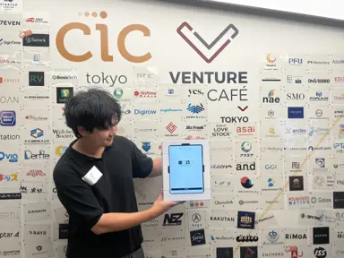 A participant holding a my4cut tablet in front of the Venture Café Tokyo sponsor wall