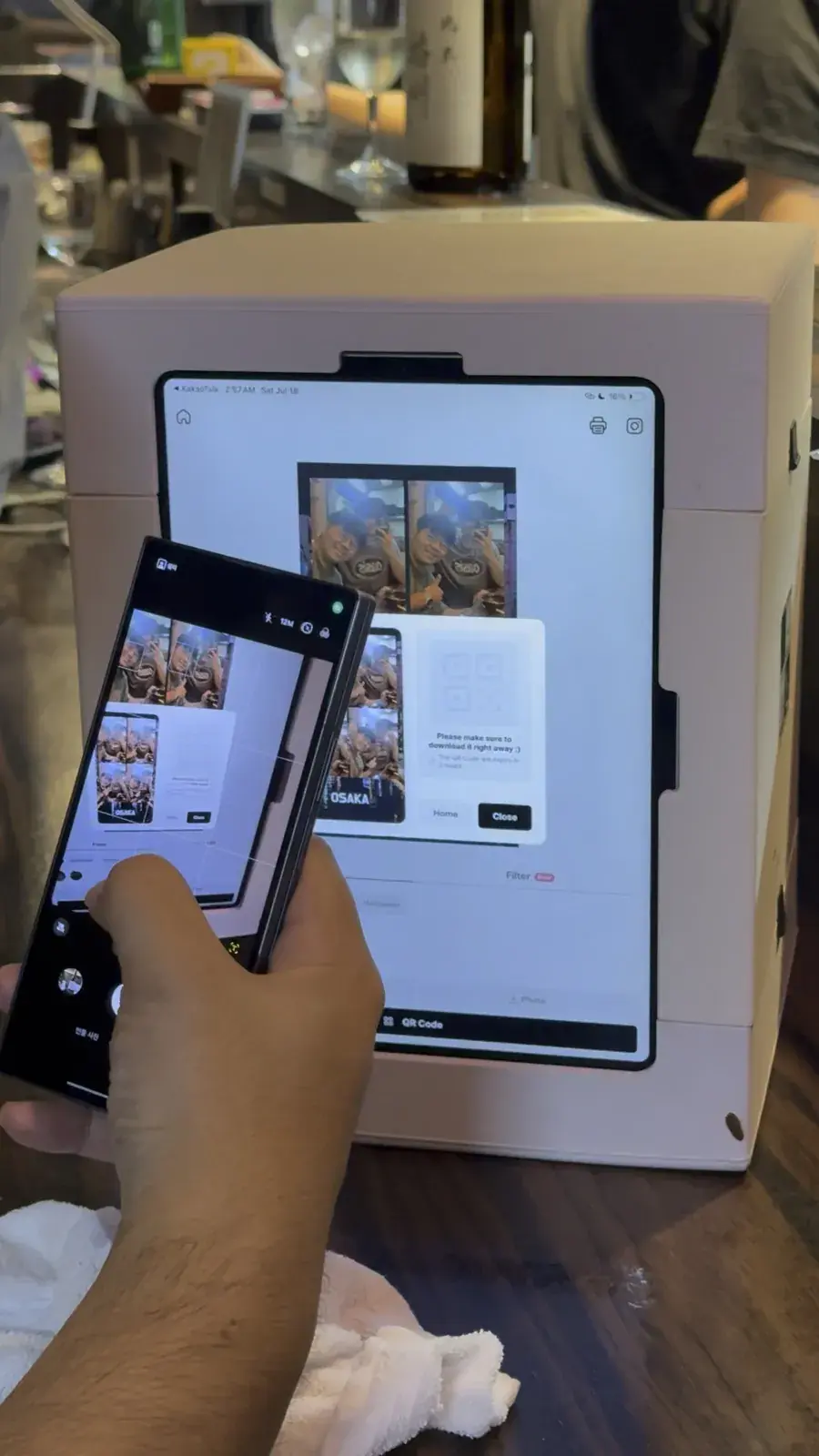 At an Osaka izakaya a hand holds a phone toward the booth screen to scan the QR code, with a four-cut strip that has the word OSAKA on screen
