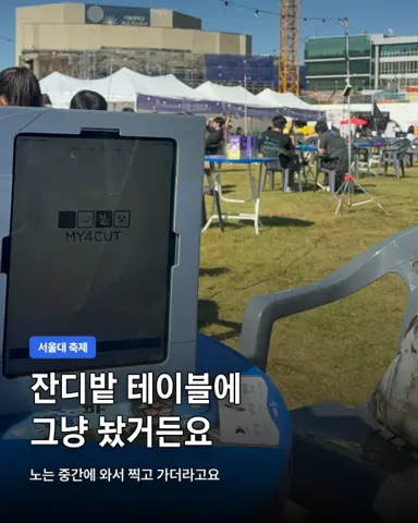 Tablet mini booth on a blue table on the Seoul National University festival lawn, MY4CUT logo on screen, white tents and people in the background