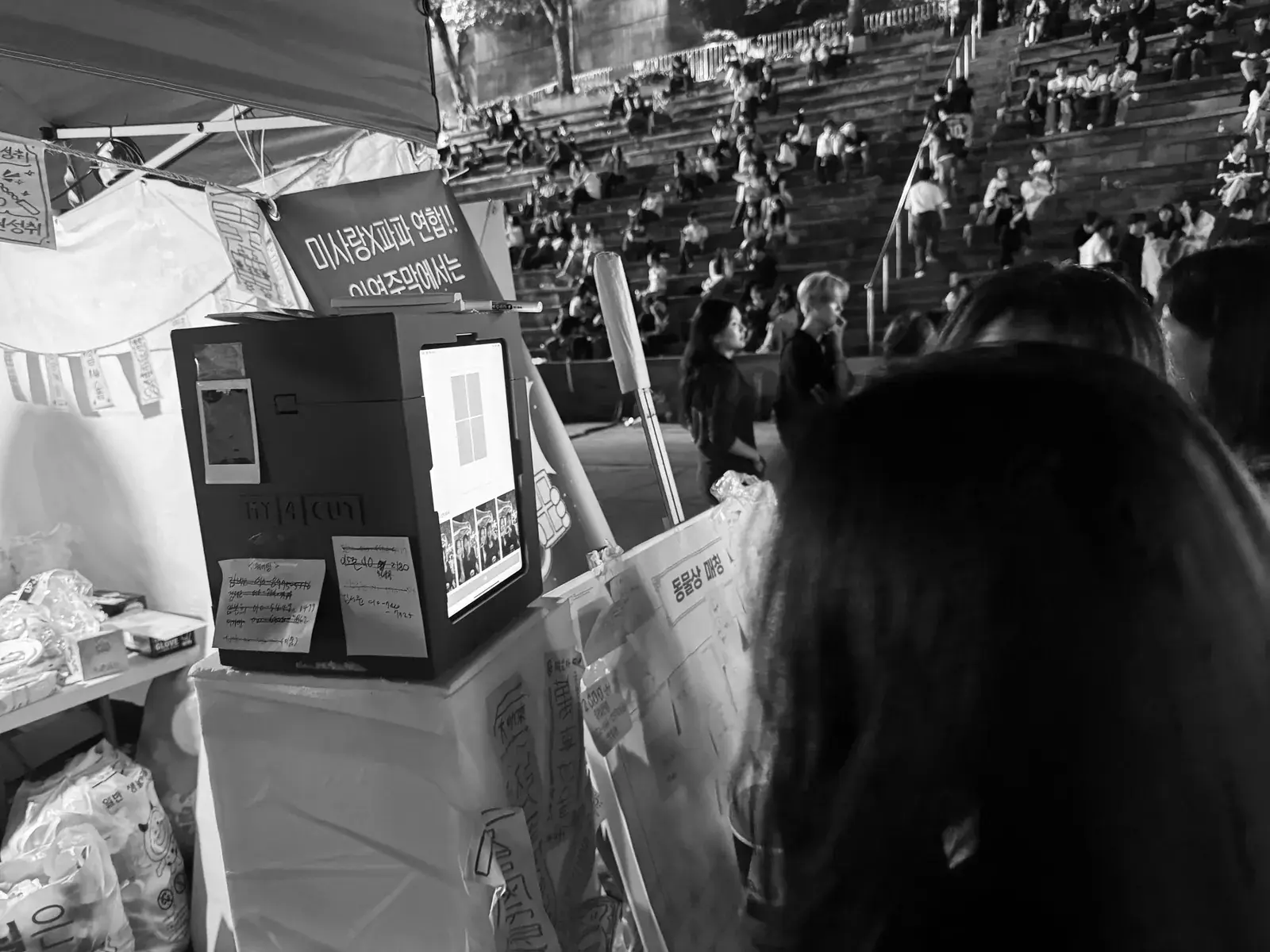 Black mini photo booth on a night festival stall beside tiered seating, a four-cut on its screen, black and white