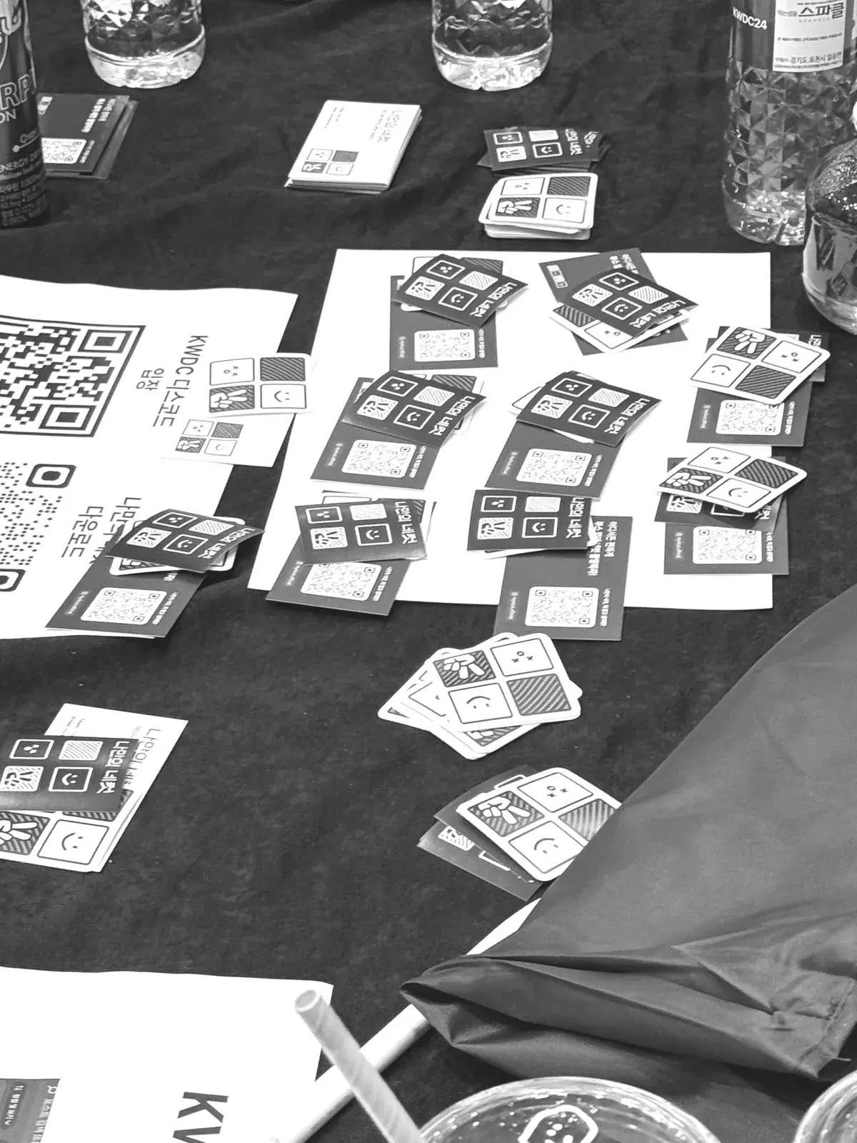Four-cut strips and QR flyers scattered on a dark event table, in black and white, with no faces visible