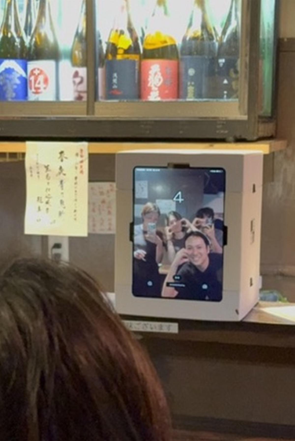 White photo booth on an Osaka izakaya counter under a shelf of sake bottles, the screen showing countdown number 4 and four people making heart poses