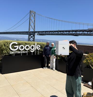 Rooftop at Google San Francisco with the Bay Bridge behind, one person holding a white mini photo booth box in the air and two people standing at the sign