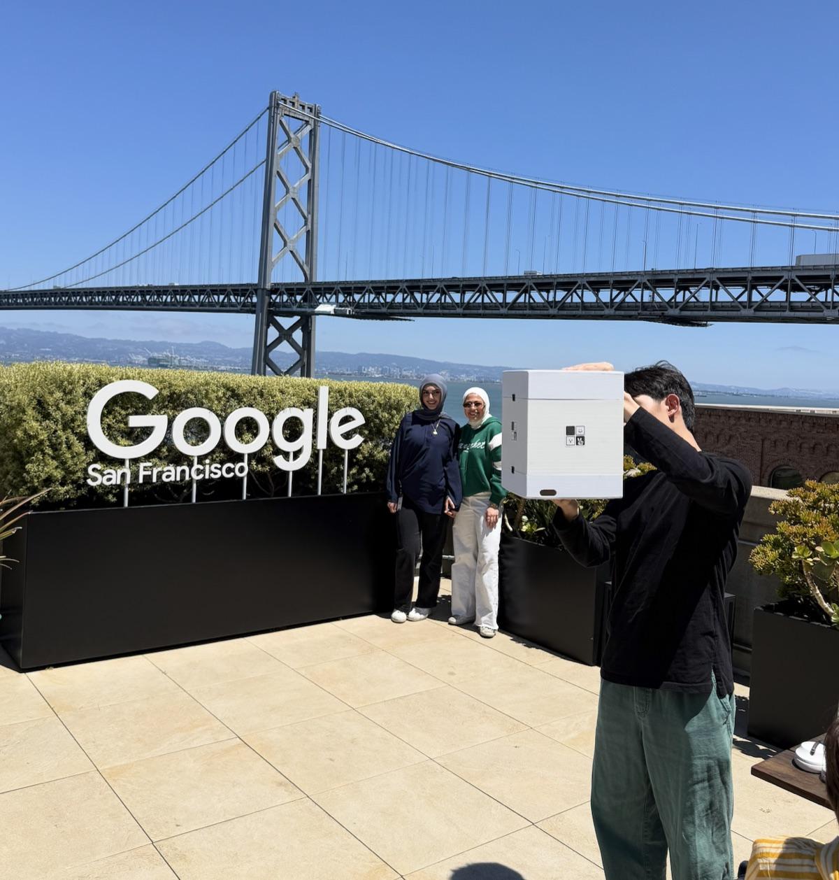 Rooftop at Google San Francisco with the Bay Bridge behind, one person holding a white mini photo booth box in the air and two people standing at the sign