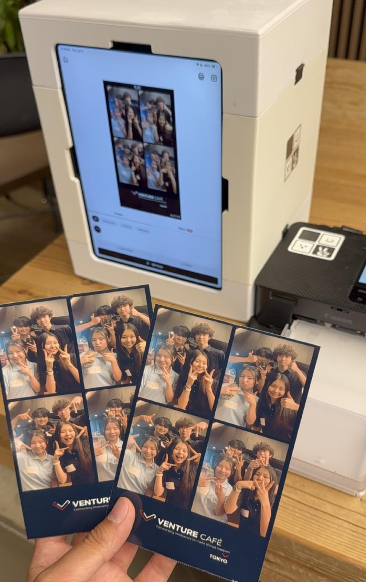 An iPad showing a four-cut result next to a Canon printer, with a hand holding two printed strips that carry the Venture Café Tokyo frame