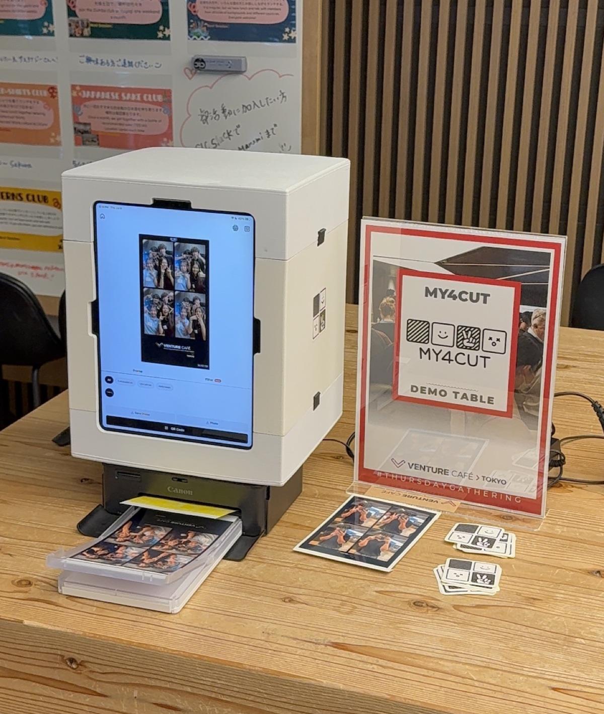 A white iPad mini booth on a Venture Café Tokyo table, a Canon printer below it with a fresh four-cut strip, and a DEMO TABLE sign beside it